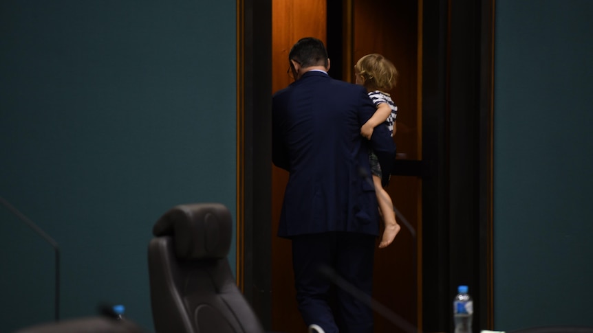 A man in a navy suit, carrying a toddler, leaving through a door in a dark green-panneled room