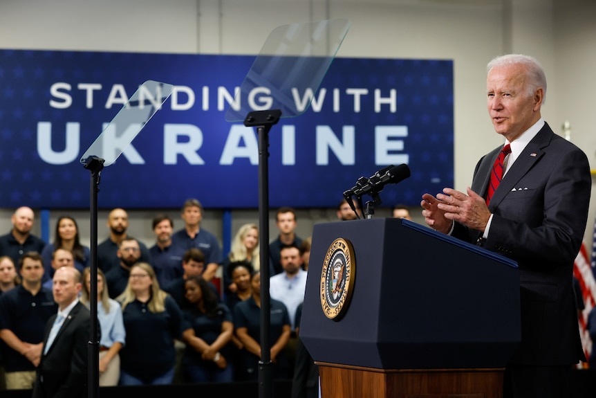Joe  Biden gives a speech in front of a sign that says 'standing with Ukraine'.