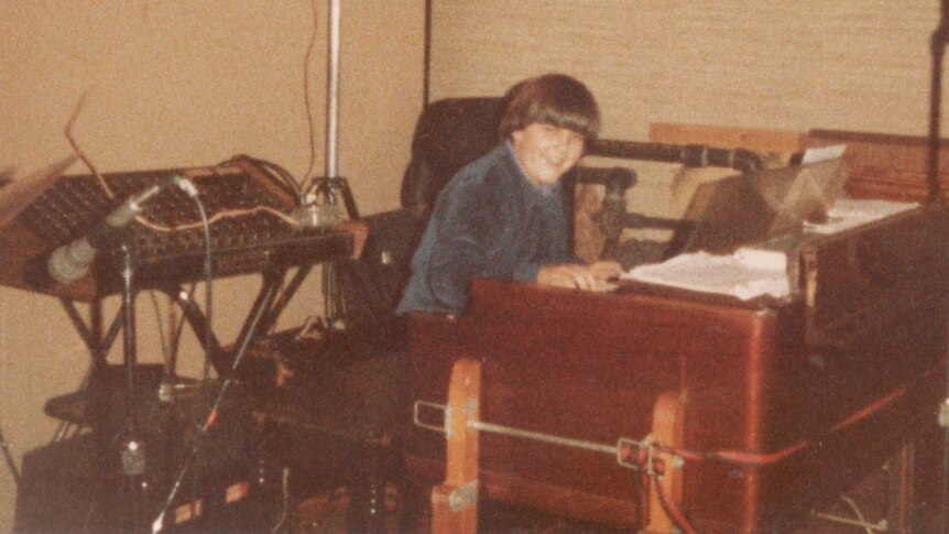 A boy with straight brown hair, smiles at the camera as he sits at an organ amid sound equipment.