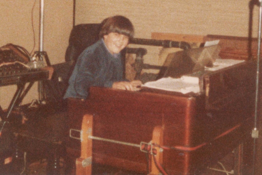 A boy with straight brown hair, smiles at the camera as he sits at an organ amid sound equipment.