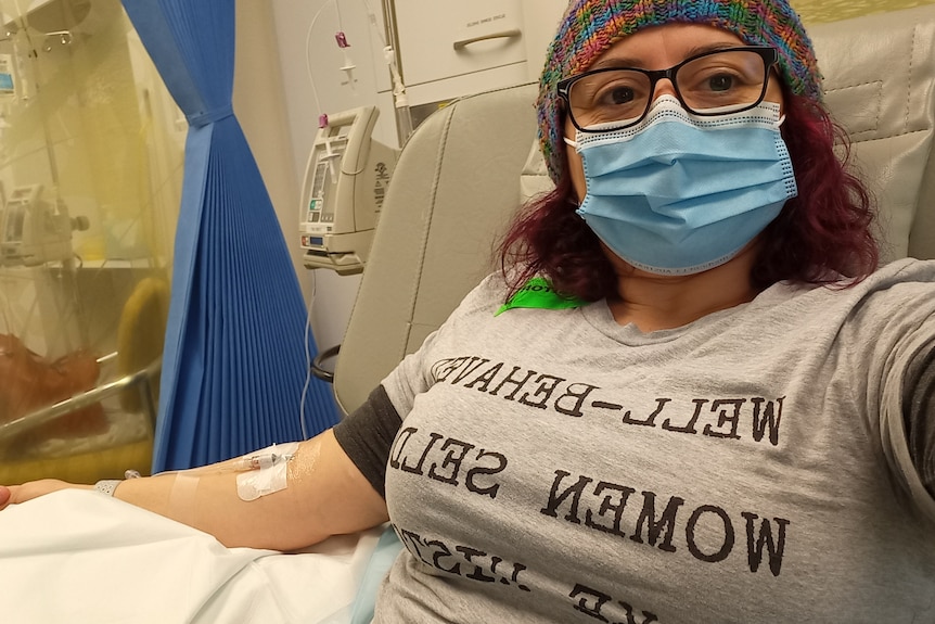 Una mujer de cabello oscuro que lleva una máscara médica recibe tratamiento intravenoso en una cama de hospital.