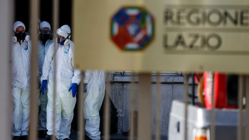 People in protective suites walk behind a gate the has "REGIONE LAZIO" written on it.