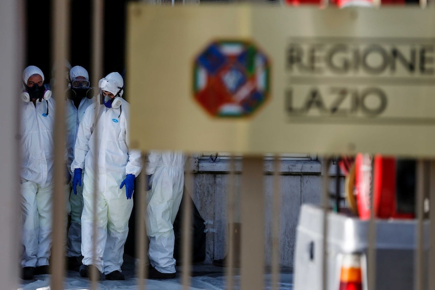 People in protective suites walk behind a gate the has "REGIONE LAZIO" written on it.