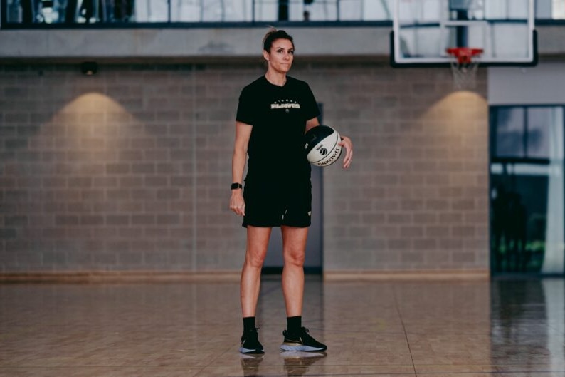 Renae Garlepp is wearing a black t-shirt and shorts, holding a ball under her arm, standing on a basketball court.