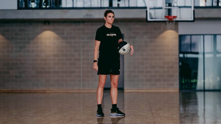 Renae Garlepp is wearing a black t-shirt and shorts, holding a ball under her arm, standing on a basketball court.