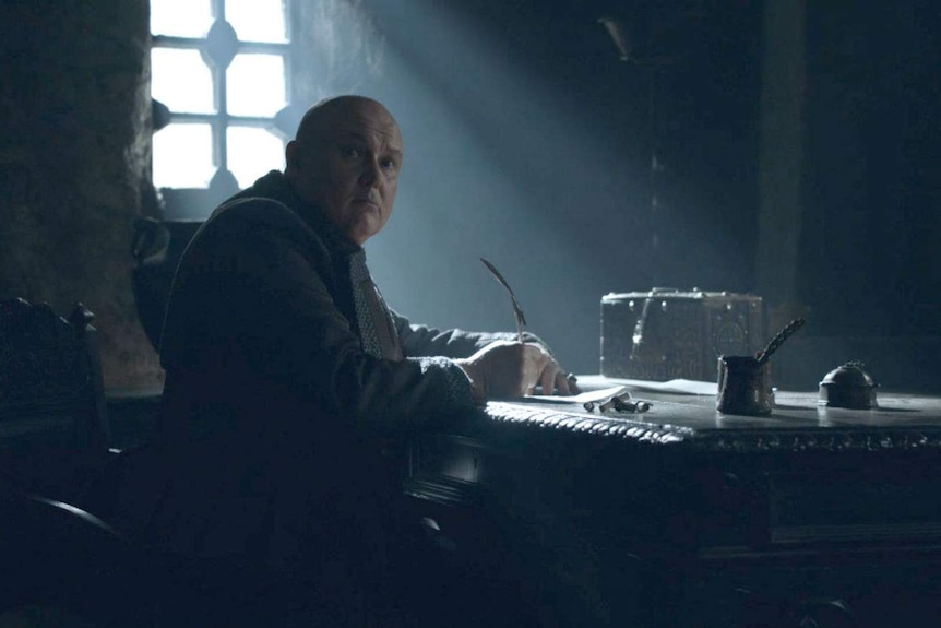 Varys writes a message at a table and looks up at someone coming.