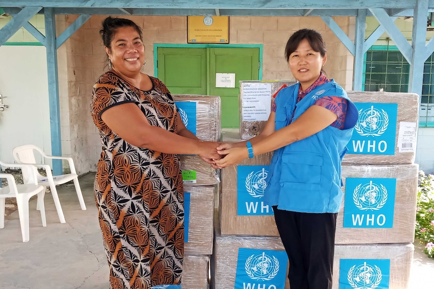 A Kiribati health service employee shakes hands with a WHO worker in front of boxes of protective equipment.