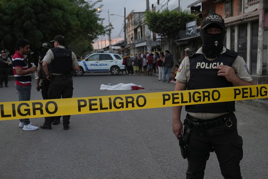 A masked police officer wearing a bulletproof vest stands on a road behind a line of yellow police tape, which reads "Peligro".