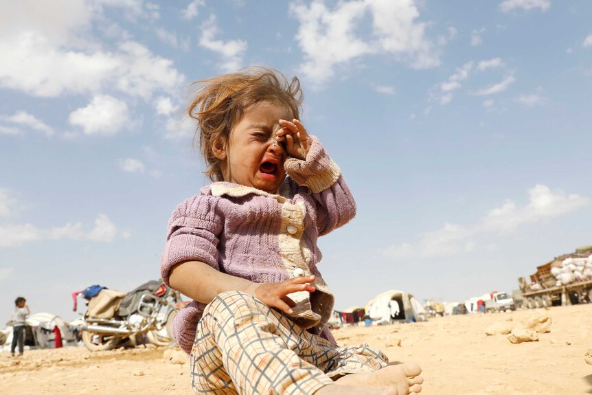 A girl wipes tears from her eyes as she cries in a dusty refugee camp.