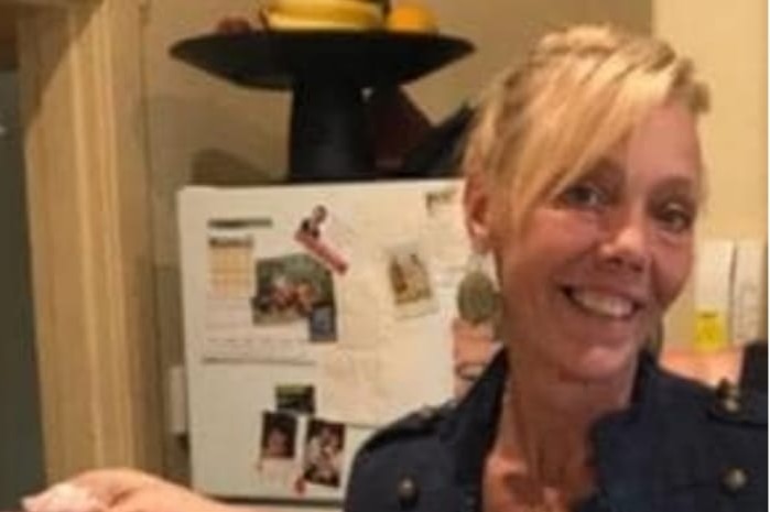 A woman with blonde hair in a kitchen holding up a pizza and smiling.