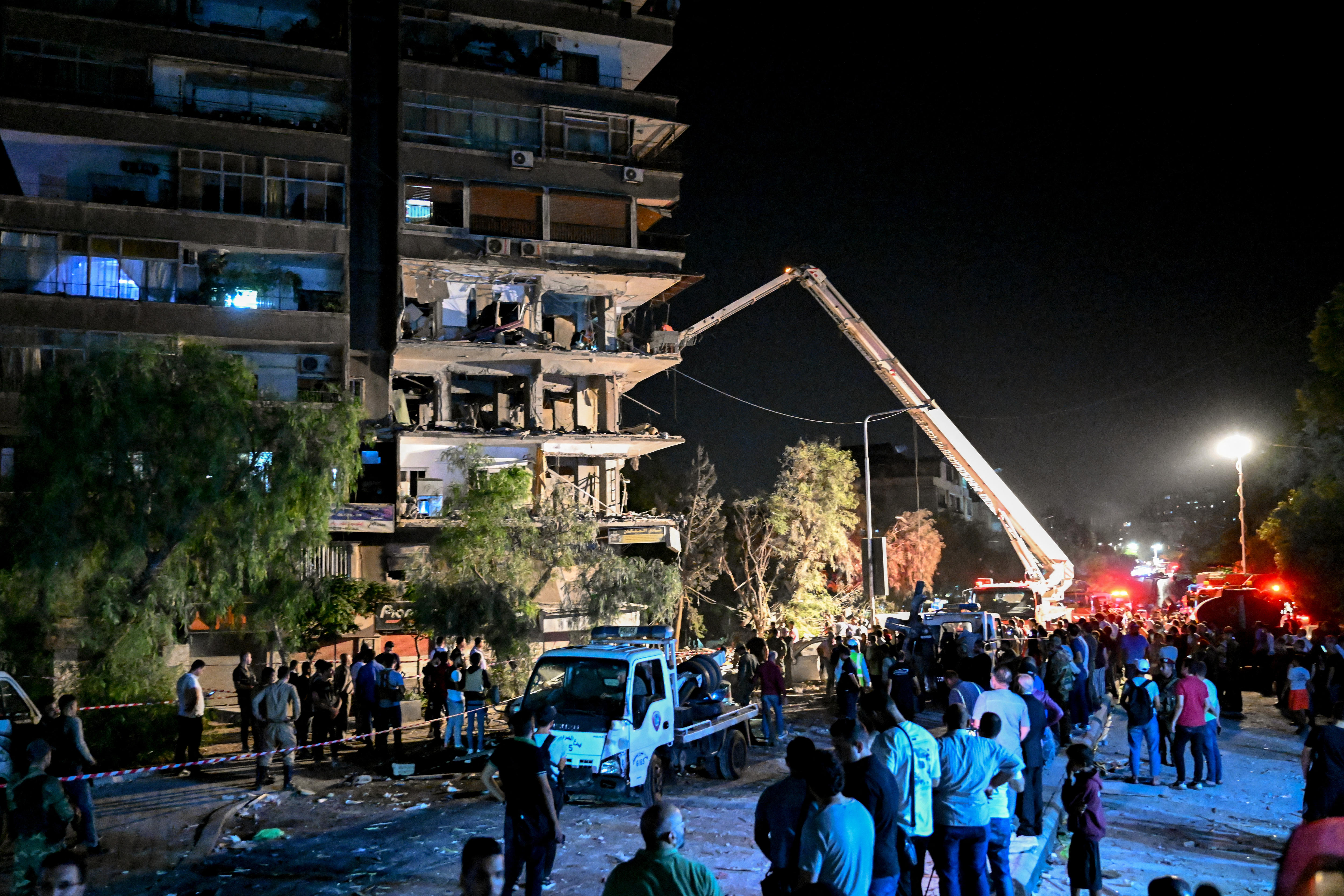 A crowd of people stand at the base of a damaged building, while a mobile crane peers into it.