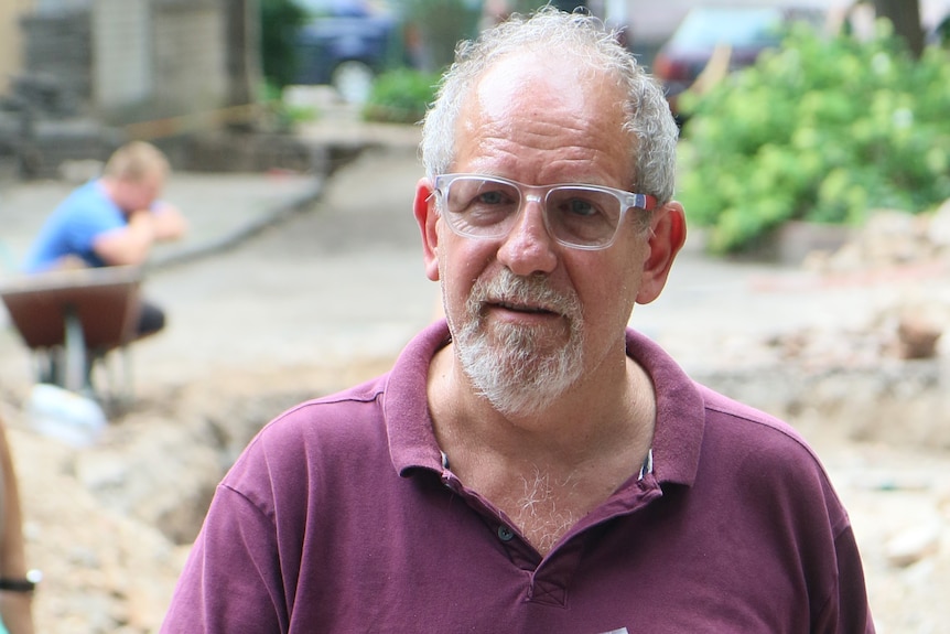 A middle-aged man with glasses and a white beard, wearing a purple polo shirt, at an excavation.