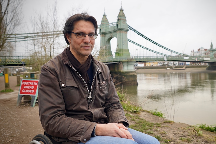 A man in a wheelchair sits in front of the closer Hammersmith Bridge.