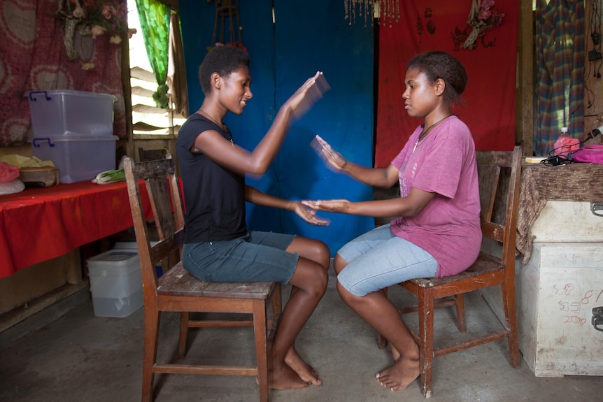 Two young Pacific girls sitting on chairs in a house learning a clapping routine.