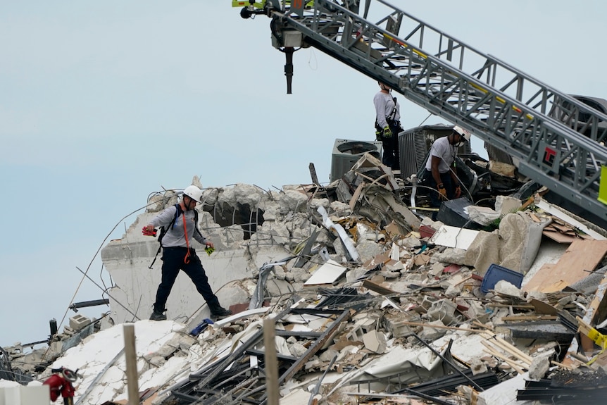 Workers in hard hats comb through rubble with a crane above them. 