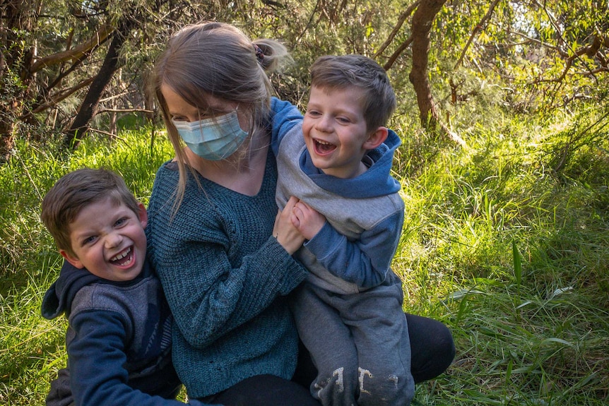 Sarah Robson, wearing a mask, hugs her two sons while standing on green grass surrounded by bush.