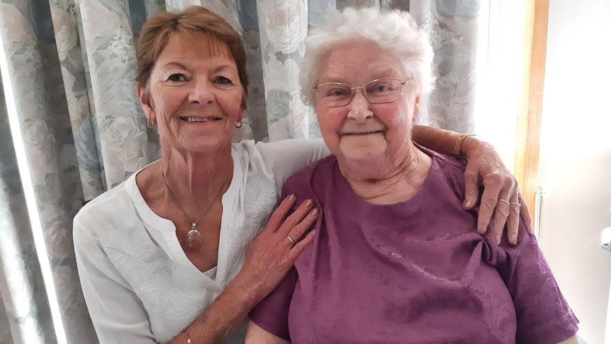 two women sit next to each other smiling, one with her arms around the other