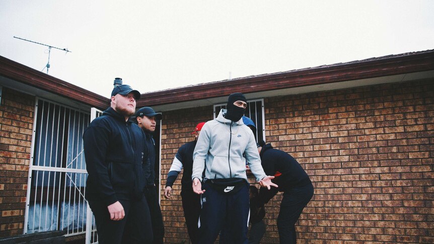 A group of men in tracksuits, one wearing a face mask, stand in front of a red brick suburban house.