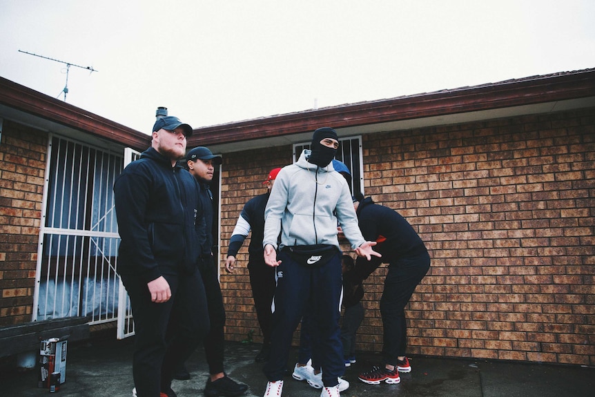 A group of men in tracksuits, one wearing a face mask, stand in front of a red brick suburban house.