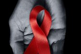 A black and white photograph of a person's hand holding a red HIV/AIDS awareness ribbon.