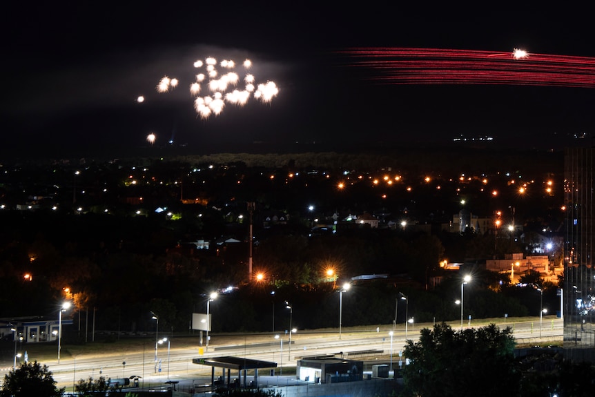 A bright cluster of white lights near a trail of red lights sits over a city a night.