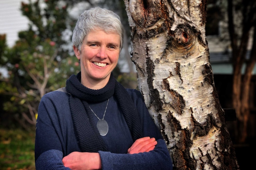 A woman with short grey hair wearing a blue top and scarf stands in front of a birch tree.