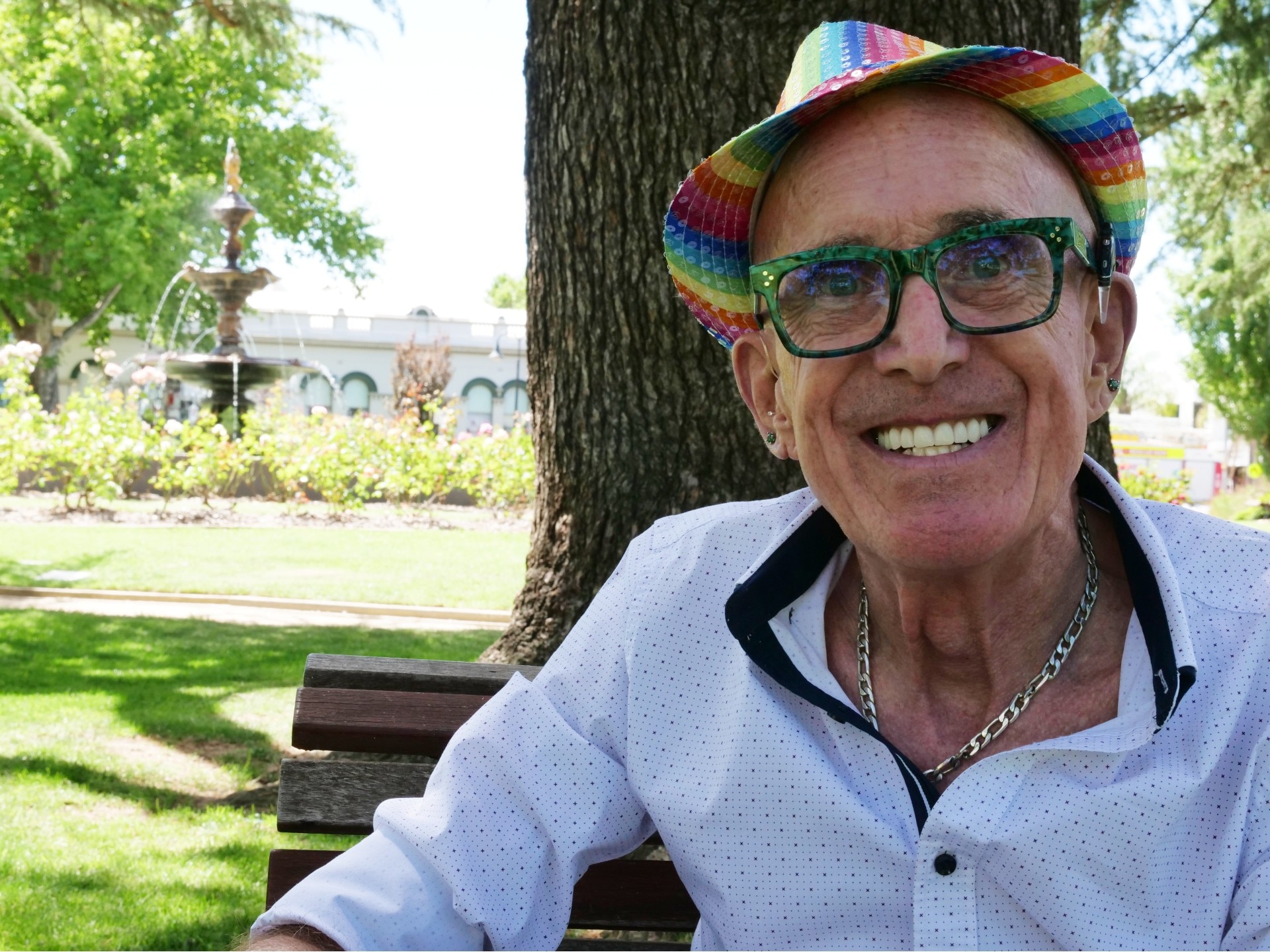 A man in a rainbow sequin hat sits in the middle of a park with a fountain behind him.