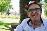 A man in a rainbow sequin hat sits in the middle of a park with a fountain behind him.