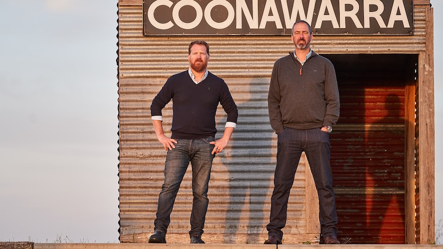 Two men stand in front of a tin shed with Coonawarra written on it. 