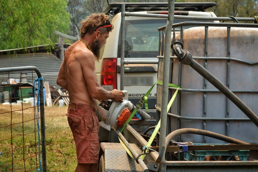A shirtless man checks a leaf blower on a trailer which has a large water tank.