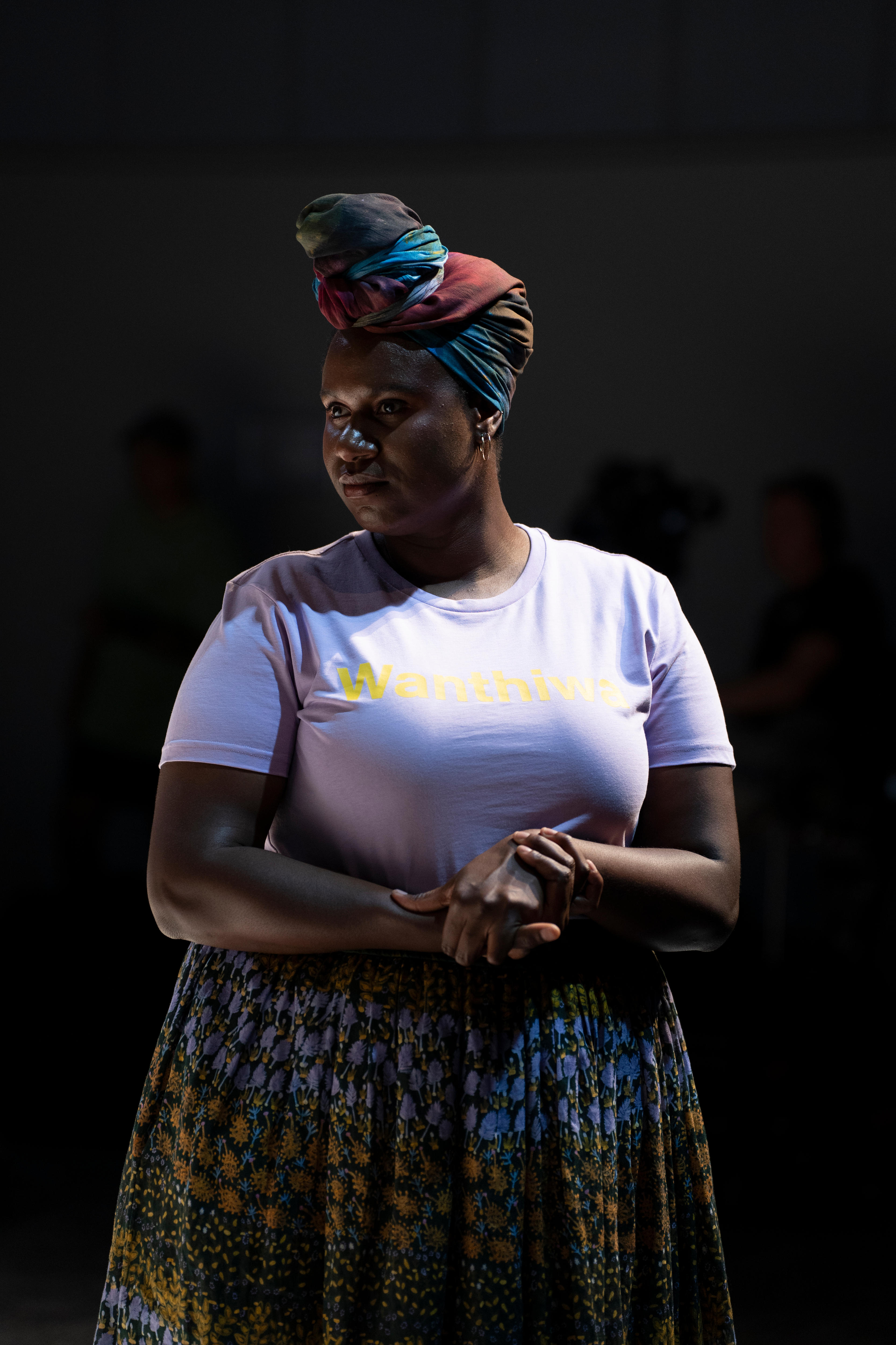 A woman wears a colorful turban and a white t-shirt with a black background.