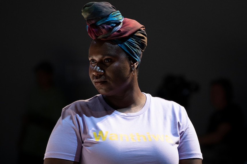 A woman wears a colorful turban and a white t-shirt with a black background.