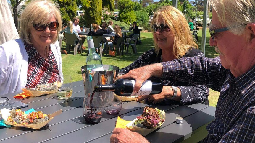 Three people sit at a table with food and wine, with lawns and other tables and guests in background