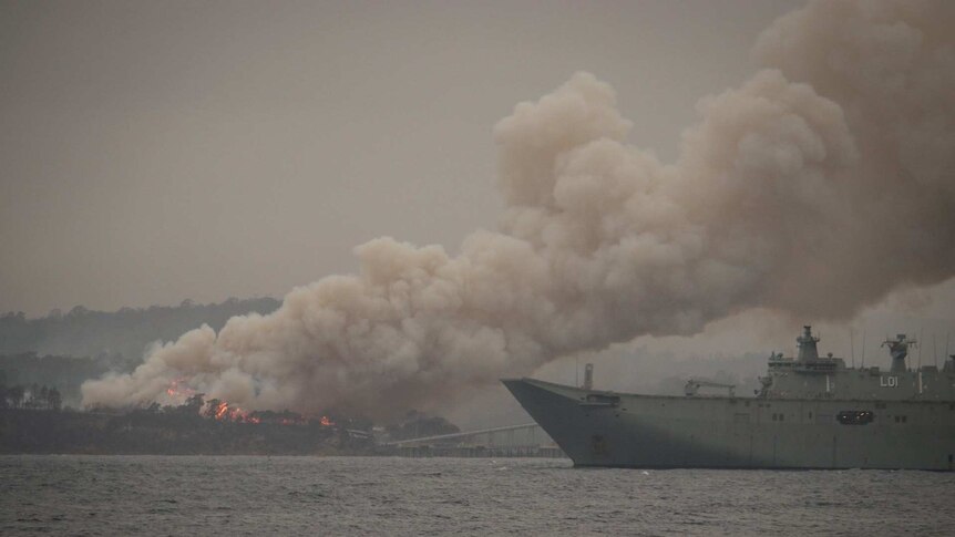 HMAS Adelaide sits off shore from Eden as the Border Fire burns in the distance