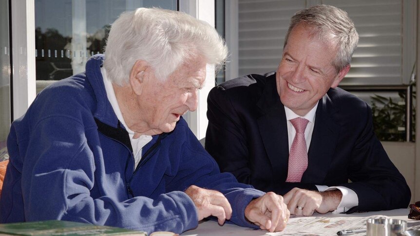 Bob Hawke (left) in a purple jumper and Bill Shorten in a suit smile while looking down at a newspaper