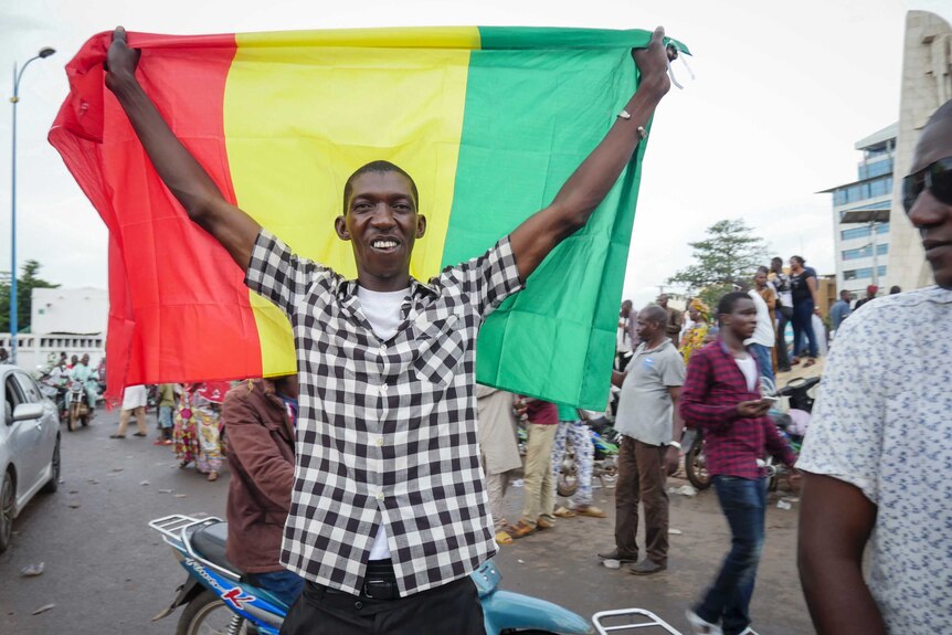 A smiling and slim black man holds a red, yellow and green flag behind on a busy African street.