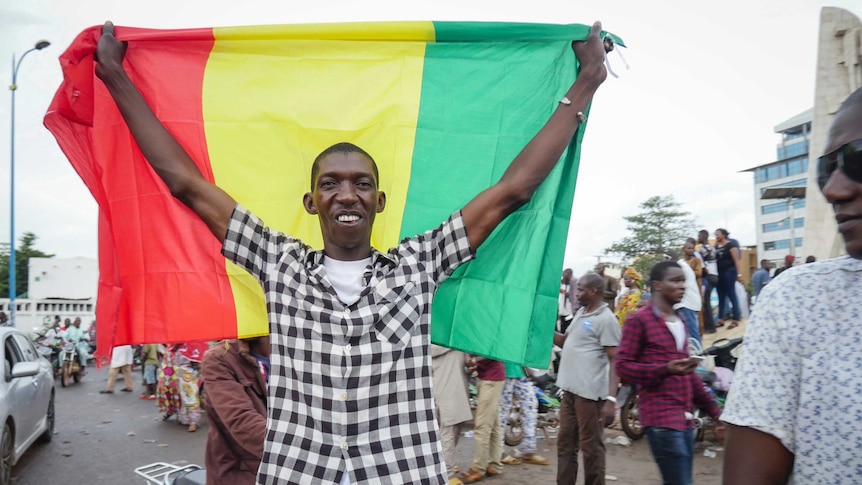 A smiling and slim black man holds a red, yellow and green flag behind on a busy African street.