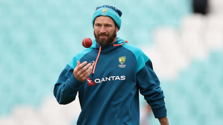 A man with a beanie and hoodie on tosses a cricket ball up in the air.