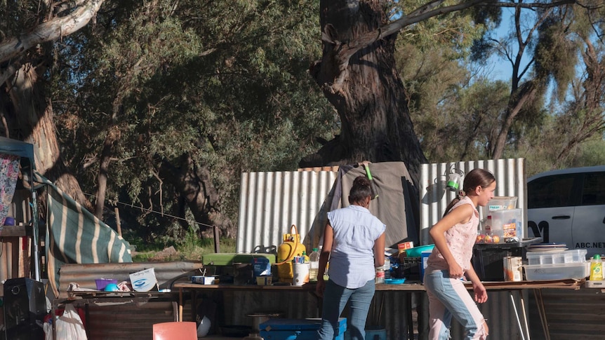 Two young women stand at an outdoor makeshift kitchen constructed of sheets of corrugated iron.