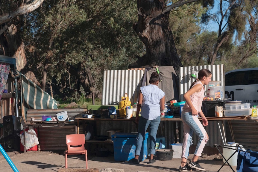 Two young women stand at an outdoor makeshift kitchen constructed of sheets of corrugated iron.