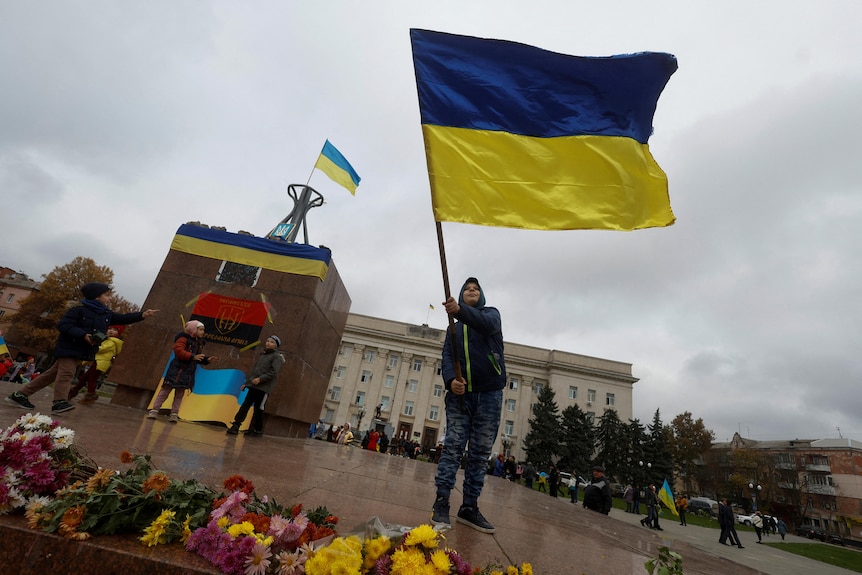 A boy waves a Ukrainian flag while standing in a town square in front of a large municipal building. 