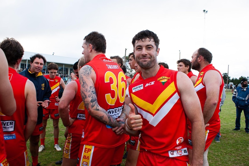 A man in a red and white and yellow guernsey smiling with a thumb up in front of a group of other men wearing the same outfit.