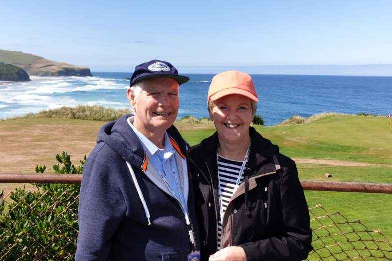 A smiling man and woman wearing caps and jackets stand together. Behind them is grass and the ocean and a bright blue sky
