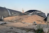 A pile of grain spills out of a badly damaged corrugated metal warehouse.