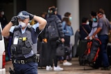 A policeman in a face mask standing a distance away from a group of people with bags outside a hotel