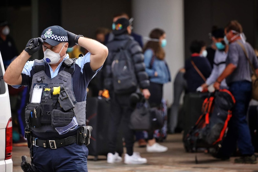 A policeman in a face mask standing a distance away from a group of people with bags outside a hotel