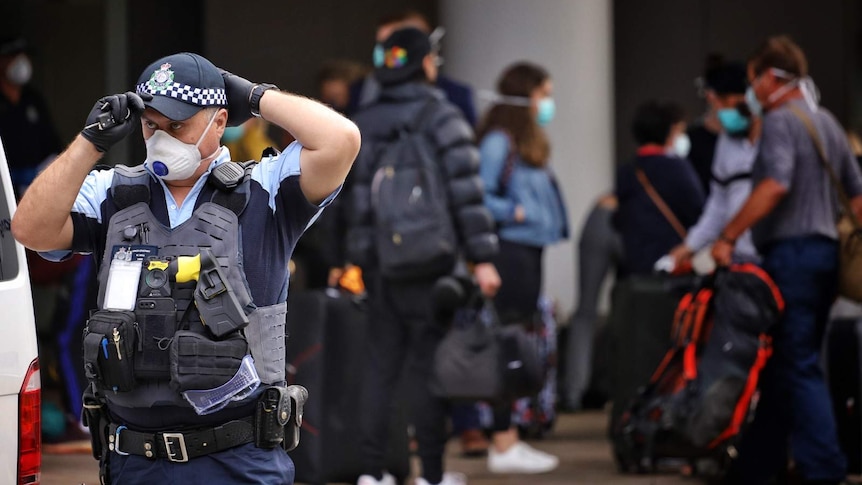 A policeman in a face mask standing a distance away from a group of people with bags outside a hotel