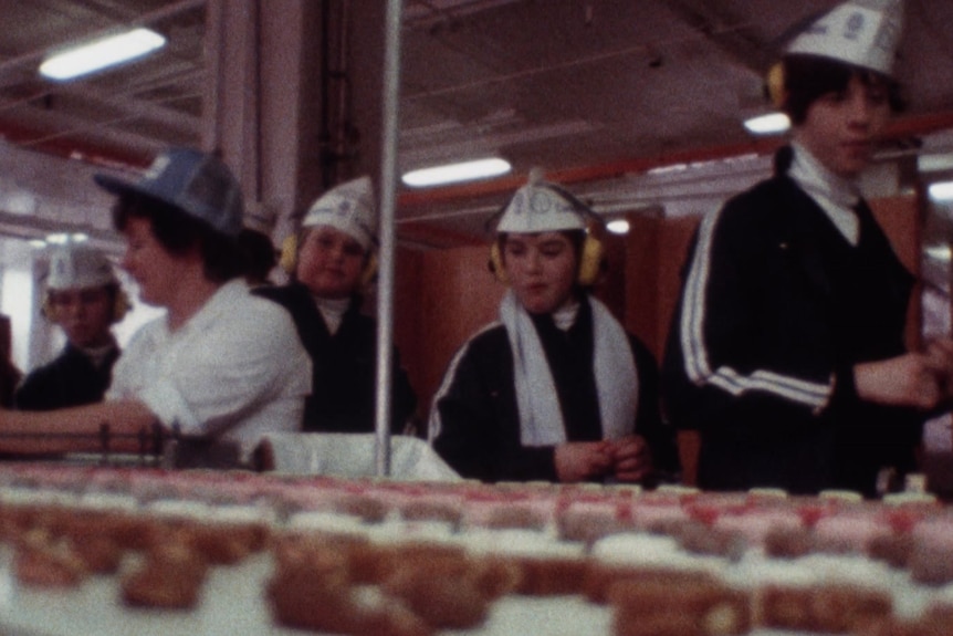 A group of children walk past a conveyor belt of chocolates, where a woman is sorting by hand