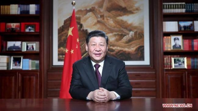 Chinese President Xi Jinping sits at a table with a bookshelf behind him.