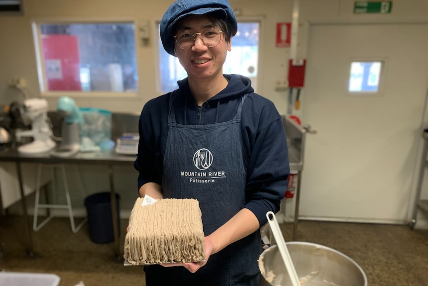 A young man in a blue apron holds a cake.